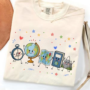 May include: A cream-colored t-shirt featuring a graphic print of a cartoon compass, globe, map, book, and mountain, all with smiling faces. The design includes colorful stars, hearts, and dots scattered across the fabric. The shirt has a Comfort Colors label.