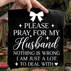 May include: Black square cushion with white text: "PLEASE PRAY FOR MY Husband NOTHING IS WRONG I AM JUST A LOT TO DEAL WITH" and a white bow graphic. The cushion is held up by a hand.