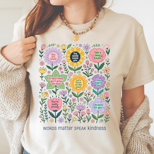 以下が含まれることがあります： クリーム色のTシャツ。カラフルな花の模様とポジティブなメッセージが特徴です。「You are loved」や「Words matter SPEAK kindness」などのフレーズが、様々な花や葉とともにデザインされています。