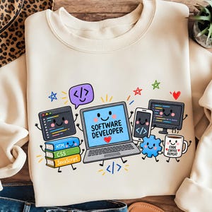 May include: A cream-colored crewneck sweatshirt featuring a colorful graphic of a smiling laptop with the text "SOFTWARE DEVELOPER" and a red heart. Surrounding elements include a stack of books labeled HTML, CSS, and JavaScript, a coffee mug with "CODE COFFEE REPEAT" text, a monitor, a smartphone, and a blue gear icon.