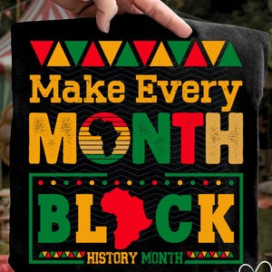 May include: Black t-shirt with the text "Make Every Month Black" in yellow, red, and green. The design includes an outline of Africa and the words "History Month". The shirt is held up by a hand.