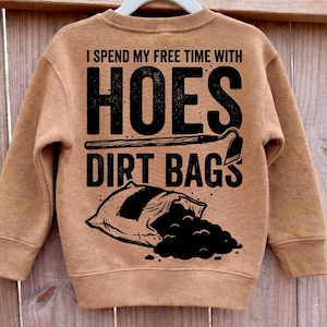 May include: A tan sweatshirt featuring the text "I spend my free time with hoes dirt bags" in black. The graphic includes a hoe, a bag of dirt, and a shovel.