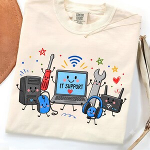 May include: A cream-coloured t-shirt featuring a colourful graphic of a laptop with the text IT SUPPORT, surrounded by cartoon-style computer hardware including a tower, mouse, router, headset, screwdriver, and wrench, all with smiling faces.
