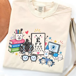 May include: A cream-coloured t-shirt featuring a colourful graphic of anthropomorphic optometry equipment, including an eye chart with letters, a phoropter, glasses, a stack of books labelled Clear Vision, Better Lives, Optometry, and a monitor.