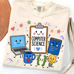 May include: A cream-colored t-shirt featuring a graphic print of a smiling clipboard with the text "COMPUTER SCIENCE" surrounded by cartoon illustrations of a laptop, a blue mug with a heart, a code window, curly brackets, and a light bulb.