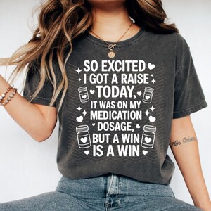 K&ouml;nnte beinhalten: Dunkelgraues T-Shirt mit wei&szlig;em Text: "SO EXCITED I GOT A RAISE TODAY, IT WAS ON MY MEDICATION DOSAGE, BUT A WIN IS A WIN." Das Design umfasst Pillenflaschen-Grafiken und Herzen.