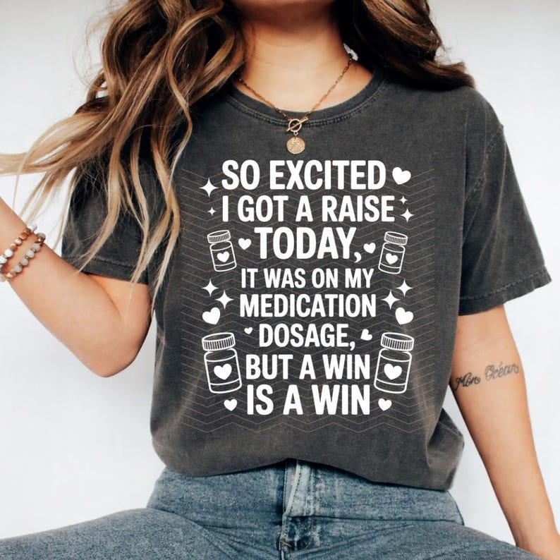 K&ouml;nnte beinhalten: Dunkelgraues T-Shirt mit wei&szlig;em Text: "SO EXCITED I GOT A RAISE TODAY, IT WAS ON MY MEDICATION DOSAGE, BUT A WIN IS A WIN." Das Design umfasst Pillenflaschen-Grafiken und Herzen.