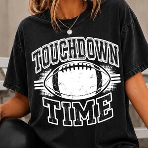 Könnte beinhalten: Ein schwarzes Kurzarm-T-Shirt mit einem abgenutzten weißen Grafikdruck auf der Brust. Das Design zeigt in der Mitte einen Football mit dem Text TOUCHDOWN darüber und TIME darunter in einer fetten Blockschrift mit verwitterter Textur.