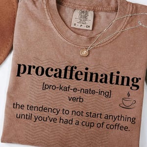 May include: A brown t-shirt with the word "procaffeinating" printed in black, along with a definition and a coffee cup graphic. The shirt has a crew neck and a small tag at the collar. A gold necklace with a pendant is draped over the shirt.