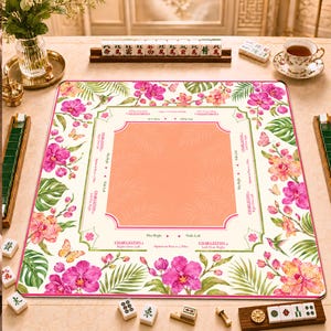 May include: A square mahjong mat featuring a peach-coloured centre with a white border decorated with pink orchids, green palm leaves, and butterflies. The border includes printed text for Charleston rules, indicating directions for passing tiles.