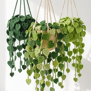 May include: Three hanging crochet plant baskets featuring trailing vines with heart-shaped leaves in various shades of green. Each basket is suspended by braided cords, showcasing intricate yarn textures and detailed leaf patterns in dark, medium, and light green tones.