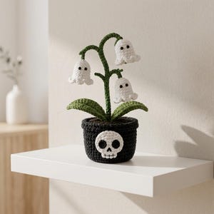 May include: A crocheted plant in a black pot featuring a white skull applique on the front. The plant has a dark green stem and leaves with three white bell-shaped flowers, each with two small black eyes, resembling ghost-like figures.