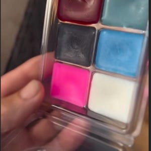 Pode incluir: Uma paleta de plástico transparente contendo seis quadrados de ceras perfumadas coloridas. As cores incluem vermelho escuro, verde-azulado, preto, azul claro, rosa choque e branco. A paleta é segurada em uma mão.