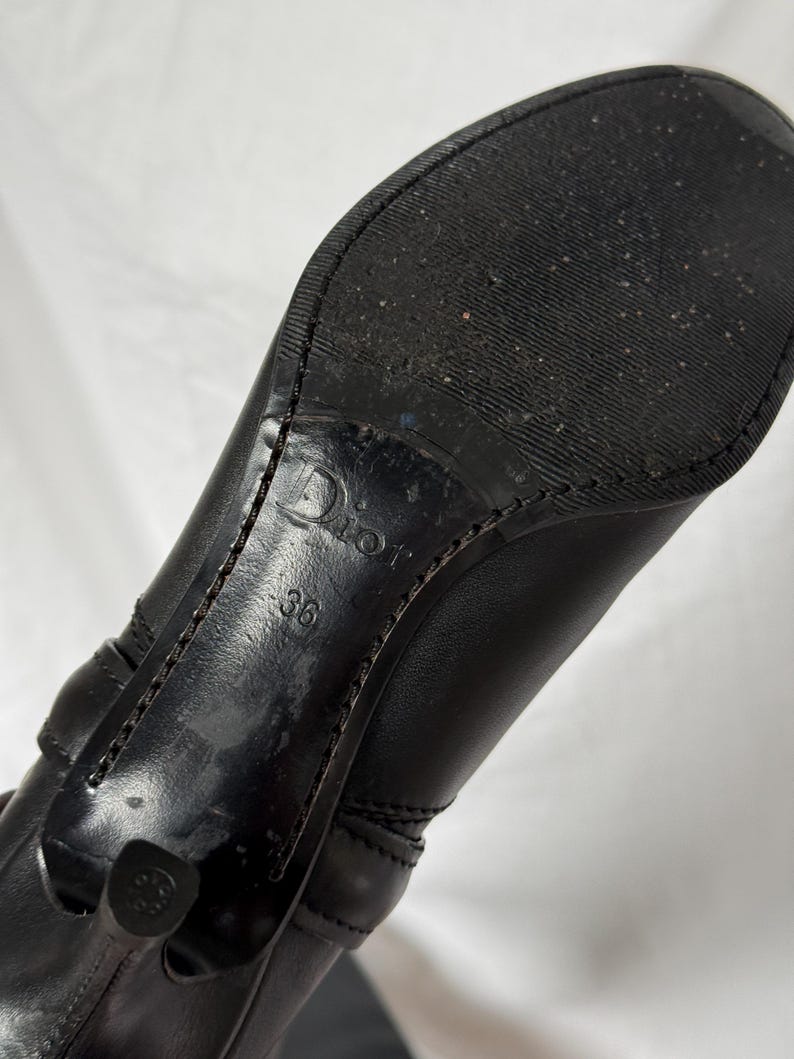 Peut inclure: Botte noire en cuir &agrave; talons hauts avec un bout carr&eacute;. La semelle est noire avec un motif textur&eacute;. Le talon est de style stiletto. La marque "Dior" est visible, ainsi que la taille "36".