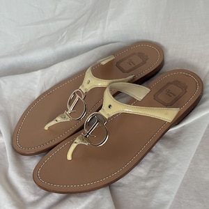 Könnte beinhalten: Ein Paar beige Leder-Zehensandalen mit einem goldfarbenen Metallakzent mit den Buchstaben "CD". Die Sandalen haben einen cremefarbenen Riemen und eine genähte Bordüre. Die Sohlen sind braun, und die Sandalen werden auf einer weißen Oberfläche präsentiert.