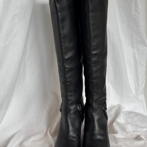 Peut inclure: Une paire de bottes noires en cuir, montant jusqu'aux genoux. Les bottes ont un bout pointu et un petit talon. Elles pr&eacute;sentent une boucle sur le c&ocirc;t&eacute;. Elles sont pr&eacute;sent&eacute;es sur un fond blanc.