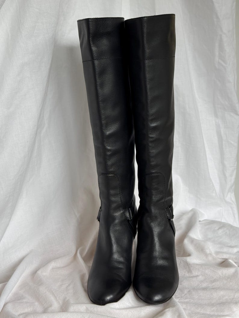 Peut inclure: Une paire de bottes noires en cuir, montant jusqu'aux genoux. Les bottes ont un bout pointu et un petit talon. Elles pr&eacute;sentent une boucle sur le c&ocirc;t&eacute;. Elles sont pr&eacute;sent&eacute;es sur un fond blanc.