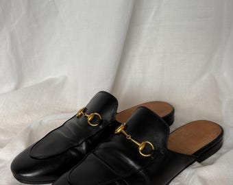 May include: A pair of black leather backless loafers with gold-colored metal horsebit detail. The shoes have a rounded toe and a leather sole. The loafers are displayed on a white background.