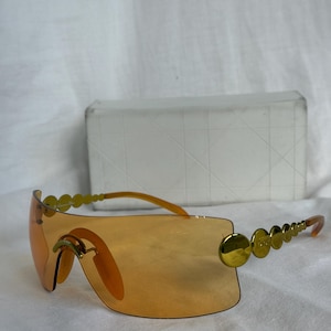 May include: Orange-tinted sunglasses with gold-colored circular accents along the top and arms. The sunglasses have a rectangular shape and a matching case in the background.