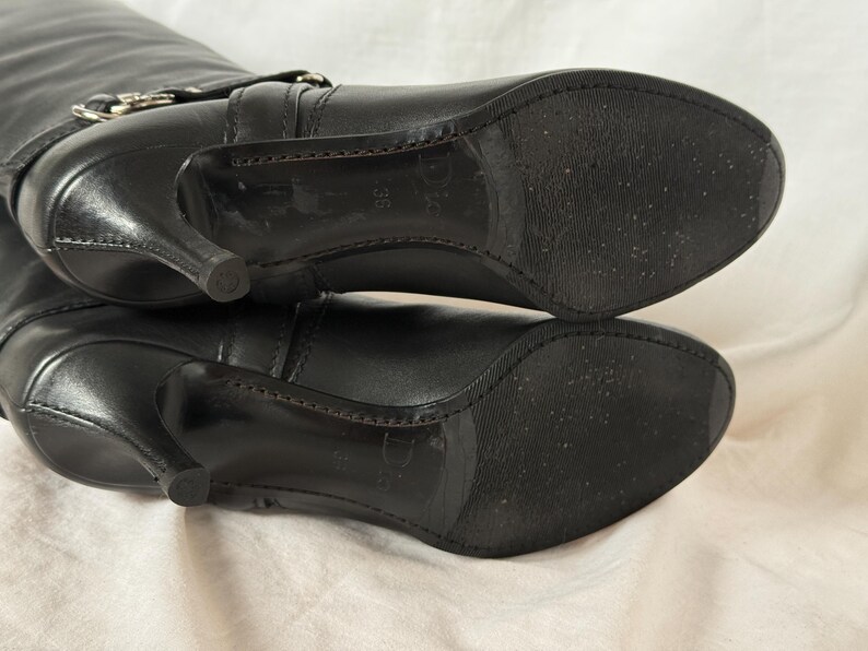 Peut inclure: Bottes hautes en cuir noir avec bout pointu et talon aiguille. Les bottes ont une boucle sur le dessus et une semelle textur&eacute;e. Les talons mesurent environ 7,6 cm de haut.