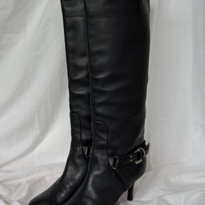 Peut inclure: Une paire de bottes noires en cuir, arrivant au genou. Les bottes ont un bout pointu, un petit talon et une boucle d&eacute;corative sur le c&ocirc;t&eacute;. Elles sont en cuir lisse et semblent en bon &eacute;tat.