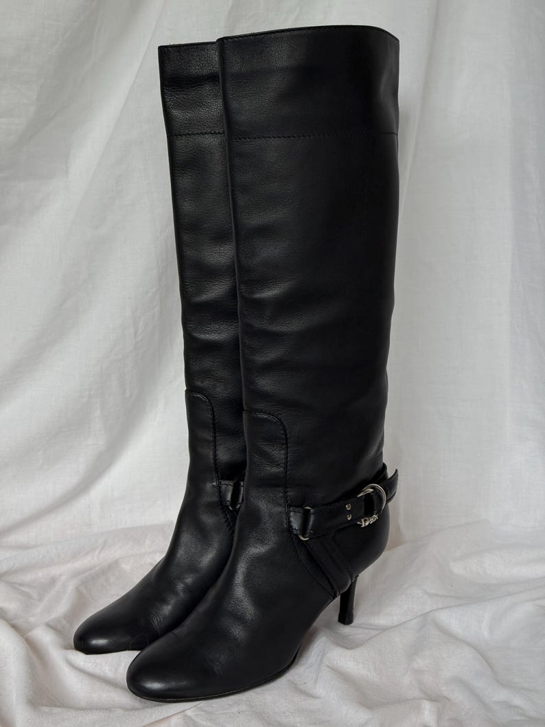 Peut inclure: Une paire de bottes noires en cuir, arrivant au genou. Les bottes ont un bout pointu, un petit talon et une boucle d&eacute;corative sur le c&ocirc;t&eacute;. Elles sont en cuir lisse et semblent en bon &eacute;tat.