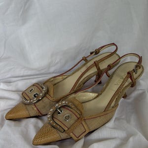 Puede incluir: Un par de tacones slingback Prada de punta. Los zapatos presentan un estampado de piel de serpiente, una hebilla decorativa con pedrería y una fina correa. Los tacones son de color marrón claro con detalles en rojo.