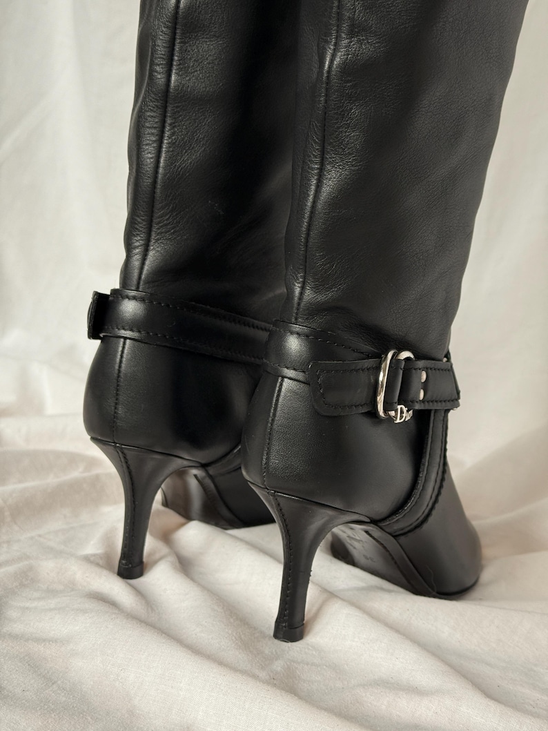 Peut inclure: Paire de bottes noires en cuir jusqu'aux genoux. Les bottes ont un bout pointu, un petit talon et une boucle sur le c&ocirc;t&eacute;. Les talons mesurent environ 7,6 cm de haut. Les bottes sont sur fond blanc.