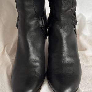 Peut inclure: Une paire de bottes en cuir noir avec un talon de hauteur moyenne. Les bottes ont une texture lisse et pr&eacute;sentent un d&eacute;tail de boucle sur le c&ocirc;t&eacute;. Les bottes sont pr&eacute;sent&eacute;es sur un fond neutre.
