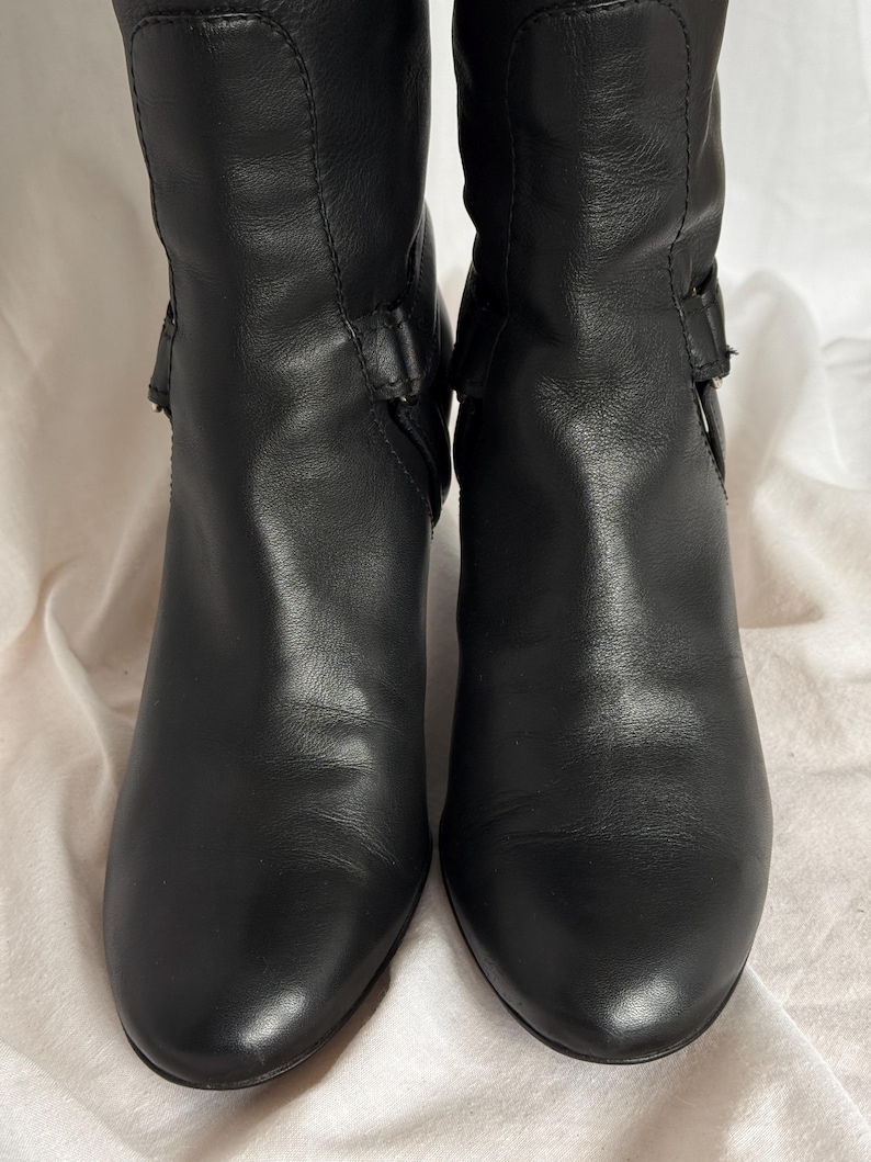 Peut inclure: Une paire de bottes en cuir noir avec un talon de hauteur moyenne. Les bottes ont une texture lisse et pr&eacute;sentent un d&eacute;tail de boucle sur le c&ocirc;t&eacute;. Les bottes sont pr&eacute;sent&eacute;es sur un fond neutre.