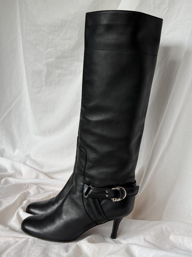 Peut inclure: Une paire de bottes hautes en cuir noir. Les bottes ont un bout pointu, un petit talon et une boucle autour de la cheville. Les bottes sont en cuir lisse et semblent en bon &eacute;tat.