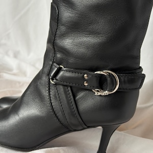 Peut inclure: Botte haute en cuir noir avec bout pointu et talon aiguille. La botte pr&eacute;sente un d&eacute;tail de bride avec une boucle argent&eacute;e et le logo Dior. La botte est en cuir lisse.