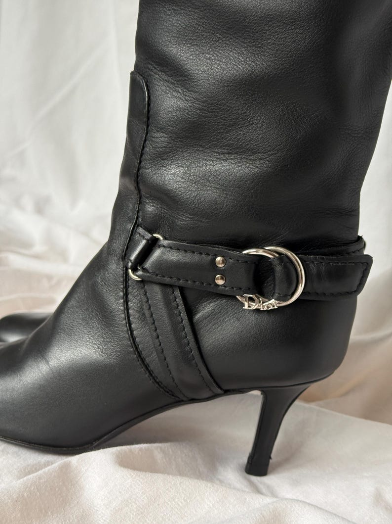 Peut inclure: Botte haute en cuir noir avec bout pointu et talon aiguille. La botte pr&eacute;sente un d&eacute;tail de bride avec une boucle argent&eacute;e et le logo Dior. La botte est en cuir lisse.