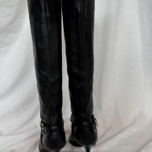 Peut inclure: Une paire de bottes noires en cuir, montant jusqu'aux genoux. Les bottes ont un bout pointu, un talon aiguille et une boucle &agrave; la cheville. Les bottes sont pr&eacute;sent&eacute;es de dos, sur fond blanc.