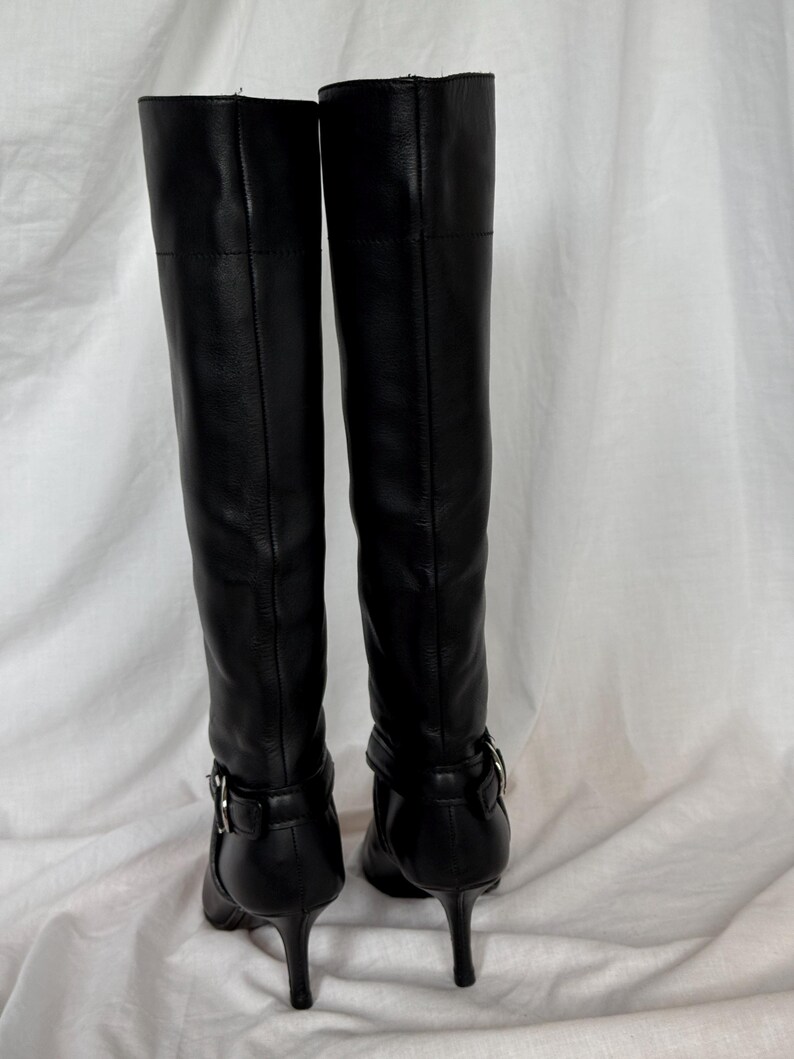 Peut inclure: Une paire de bottes noires en cuir, montant jusqu'aux genoux. Les bottes ont un bout pointu, un talon aiguille et une boucle &agrave; la cheville. Les bottes sont pr&eacute;sent&eacute;es de dos, sur fond blanc.