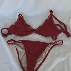 May include: A burgundy bikini with a triangle top and a high-cut bottom. The bikini has a delicate white trim.