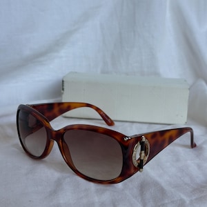 May include: Tortoiseshell sunglasses with brown gradient lenses. The oval frames have a gold-tone accent on the temples. The sunglasses are set against a white background, with a white box in the background.