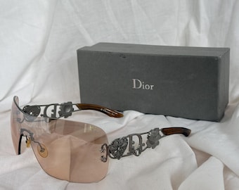 May include: A pair of Dior sunglasses with a light brown tint and decorative metal accents. The sunglasses have a curved, shield-like shape and brown arms. A gray box with the Dior logo is in the background.