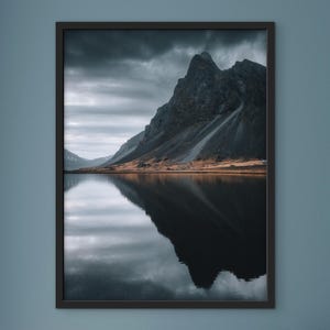 May include: A framed landscape photograph featuring a mountain reflected in a calm lake. The mountain is dark gray and the sky is overcast. The water is a dark, reflective surface. The frame is black.