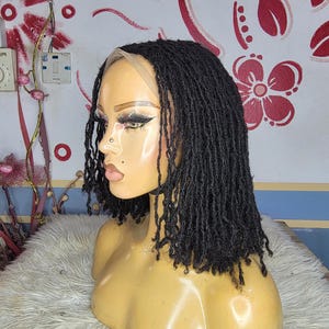 Könnte beinhalten: Schwarze Dreadlock-Perücke im Bob-Schnitt, präsentiert auf einem Schaufensterpuppenkopf. Die Perücke hat einzelne Dreadlocks und eine Lace-Front. Die Schaufensterpuppe steht auf einer flauschigen, weißen Oberfläche.
