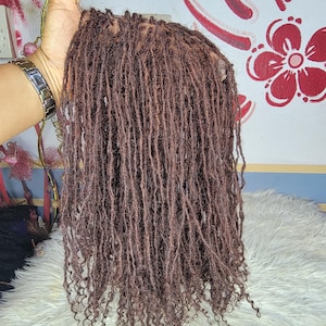 以下が含まれることがあります： 多数の細いテクスチャー加工されたドレッドヘアがレイヤードスタイルで垂れ下がるブラウンのウィッグ。髪の毛はわずかに縮れた自然な質感で、毛先はカールしており、ウィッグ上部にはメッシュキャップのベースが見えています。