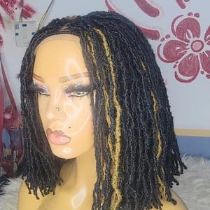 Può includere: Una testa di manichino che mostra una parrucca di dreadlocks neri e biondi. La parrucca presenta dreadlocks corti e strutturati con ciocche bionde. Il manichino ha il trucco applicato, tra cui ombretto e rossetto. Lo sfondo è un muro con motivi decorativi.