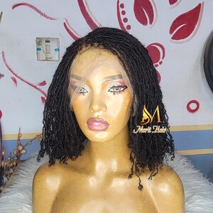 Peut inclure: Perruque noire avec micro-tresses, coiffée en carré court. La perruque présente un devant en dentelle et est présentée sur une tête de mannequin. Le mannequin est maquillé, avec du rouge à lèvres rose et de l'ombre à paupières. Le texte "Merit Hair" est visible.