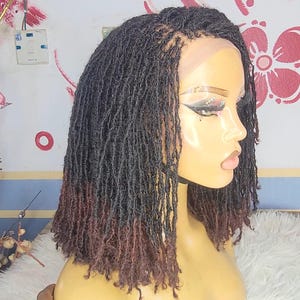 May include: A mannequin head displaying a short, black wig with reddish-brown tips. The wig features small, individual dreadlocks. The mannequin has makeup applied, including eyeshadow and lipstick. The head is set against a patterned wall.
