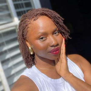 May include: A person with brown locs and gold hoop earrings. They are wearing a white tank top and a delicate gold necklace. The person is touching their face with their right hand, and their lips are painted a deep red.