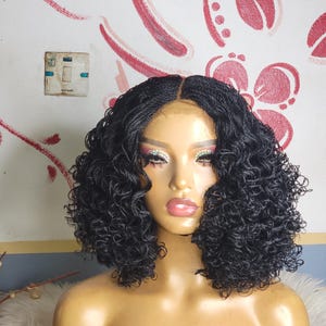 May include: A black, curly wig with a center part is displayed on a mannequin head. The wig features small, tightly woven braids that create a voluminous, textured look. The mannequin has full makeup, including bold eye shadow and lipstick.