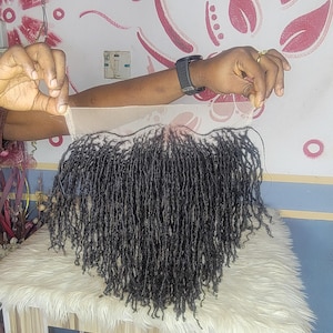 May include: A black wig with micro locs, featuring a lace front. The wig is displayed on a white, fluffy surface. The hair is a dark shade, and the lace front is held up by a person's hands.