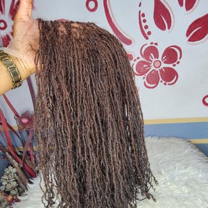May include: A bundle of brown micro locs, held up against a patterned background. The locs are tightly coiled and have a natural, textured appearance. The hair appears to be a dark brown color.