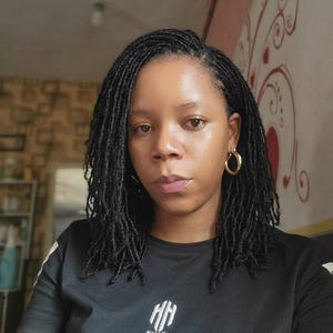 May include: A person with black locs and gold hoop earrings. The person is wearing a black t-shirt with white stripes on the shoulders and a white logo that says "KHAN" at the bottom.