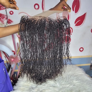 May include: A dark brown wig with long, thin, curly strands. The wig is held up by a hand, showing the lace front and the texture of the hair. The hair appears to be a natural colour with some reddish highlights.